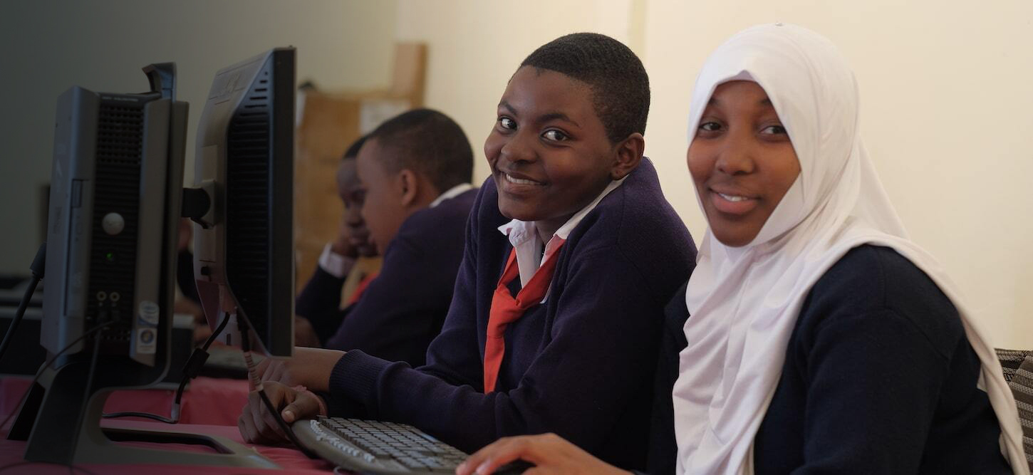 two high school students sitting in front of computers in a classroom smiling at camera