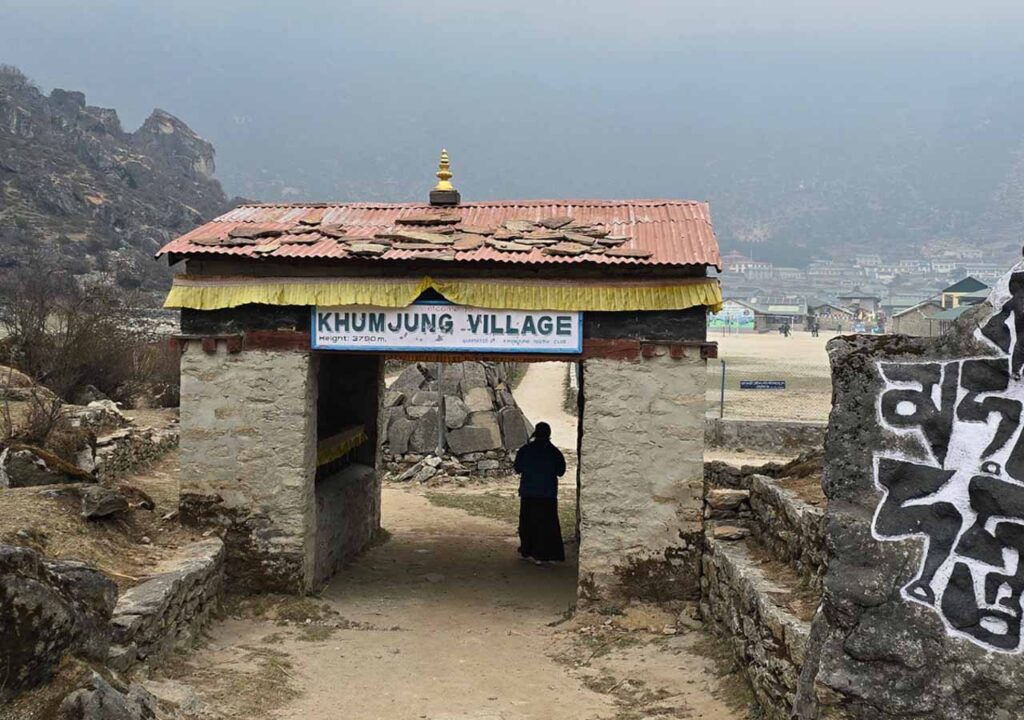 A sign reads 'Khumjung Village' on a stone arch entryway.