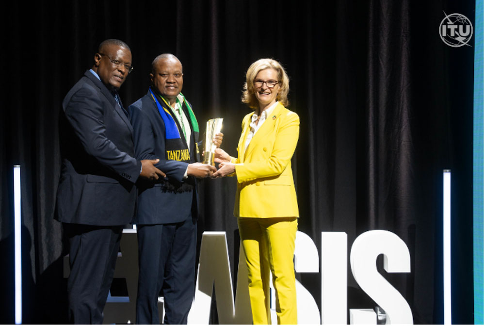 Two men—one wearing a scarf that says 'Tanzania'—accept an award from a woman.