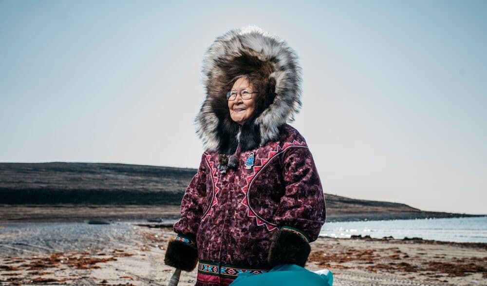 An indigenous woman stands outdoors in a fur-hooded coat with water in the background.