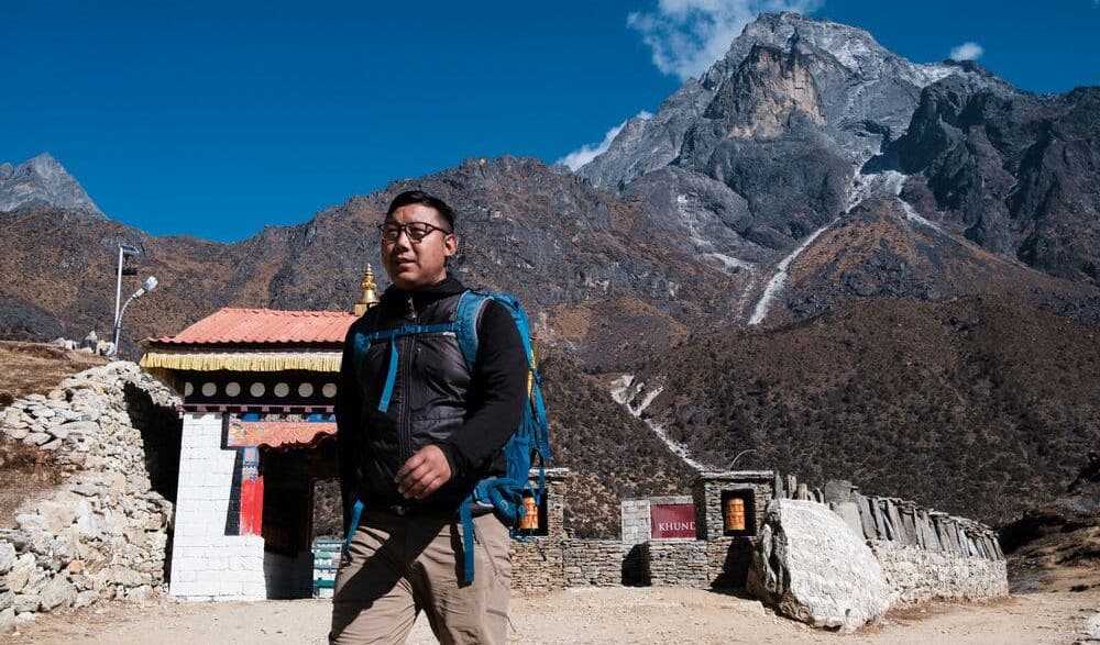 A man walks through a village with a mountain in the background.