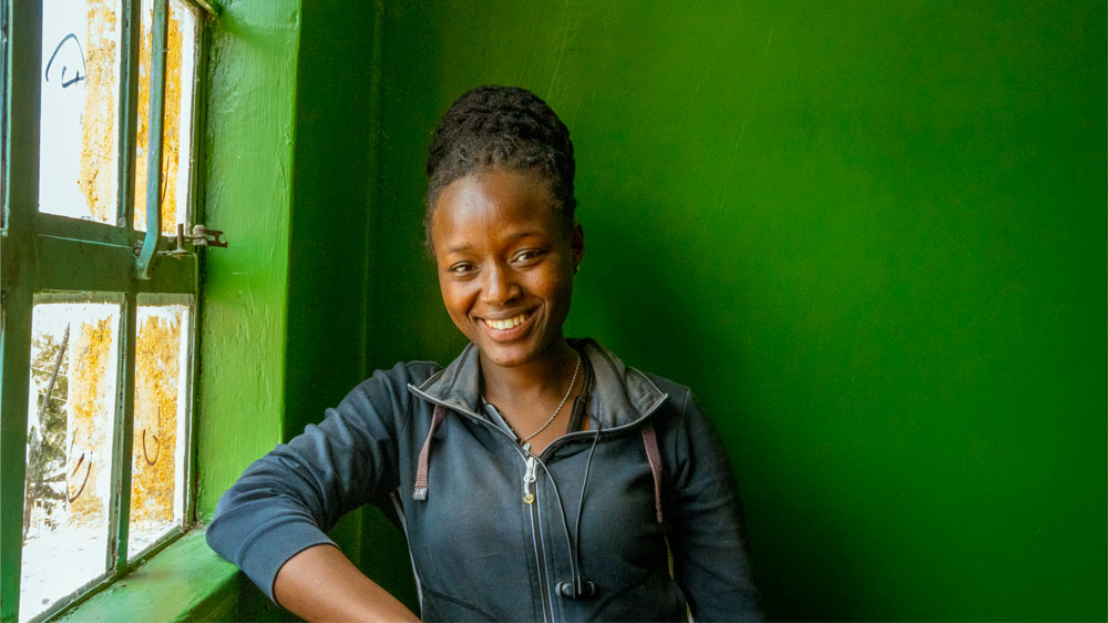 Ruth Njeri stands and smiles in front of a green wall